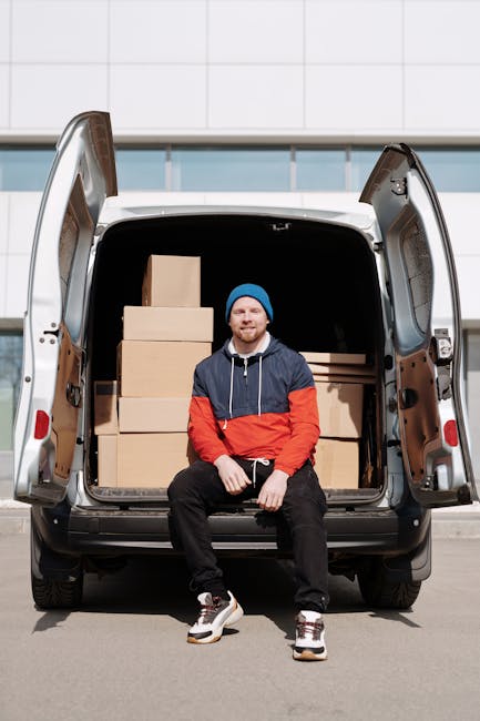 A young man with a beard, wearing a blue knit cap, a navy and red jacket, black trousers, and white sneakers, is sitting on the edge of an open van's rear loading area. Inside the van, there are sever