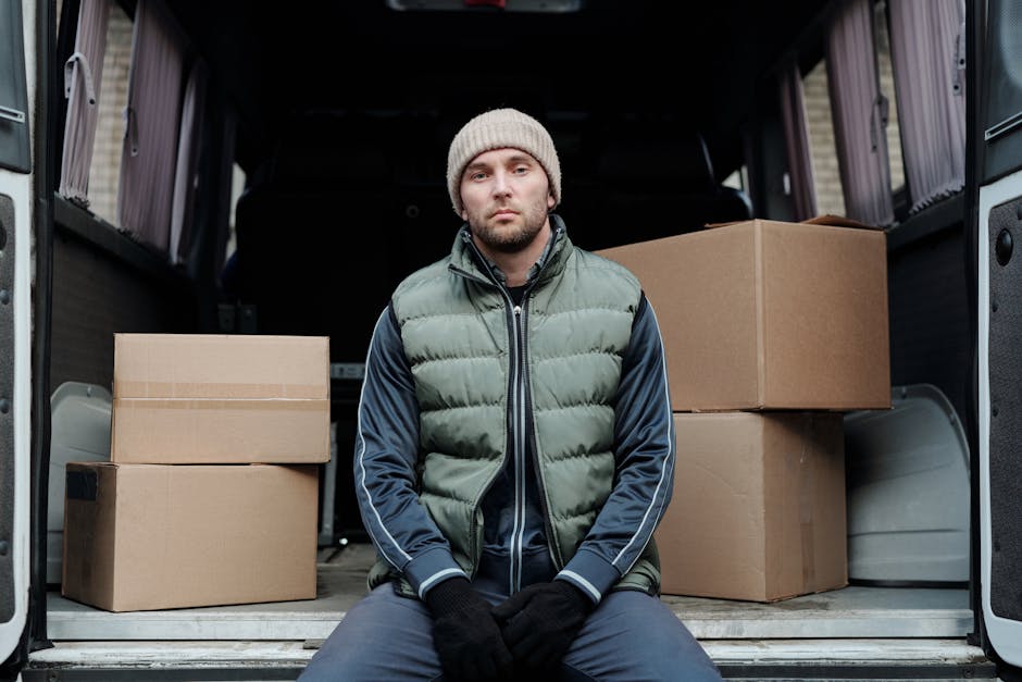 A young man with a beard, wearing a blue knit cap, a navy and red jacket, black trousers, and white sneakers, is sitting on the edge of an open van's rear loading area. Inside the van, there are sever