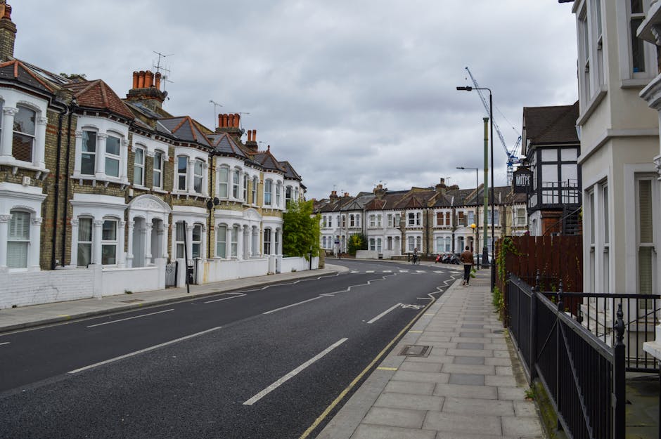 A narrow residential street in West Norwood SE27 with a brick-paved surface, lined with closely spaced terraced houses on both sides, some with white or brick facades and small front gardens filled wi